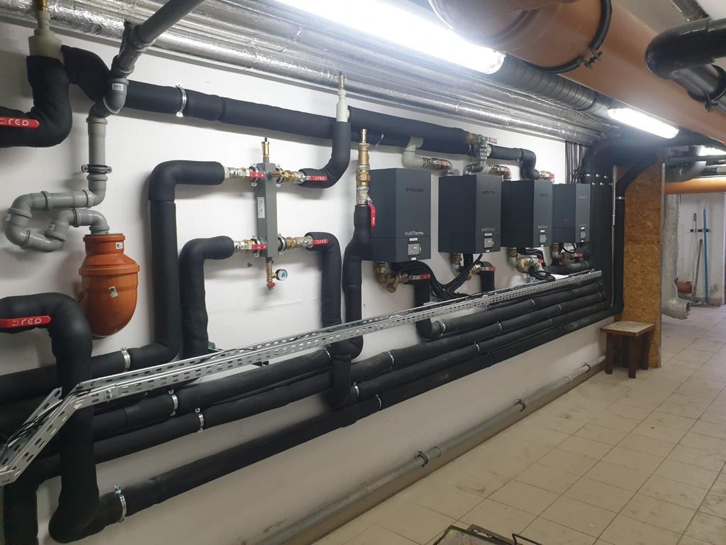 Piwnica z czterema czarnymi kotłami grzewczymi MultiThermo zamontowanymi na białej ścianie, połączonymi rurami w czarnej izolacji, z pomarańczowym filtrem, na tle kafelkowej podłogi i drewnianej...