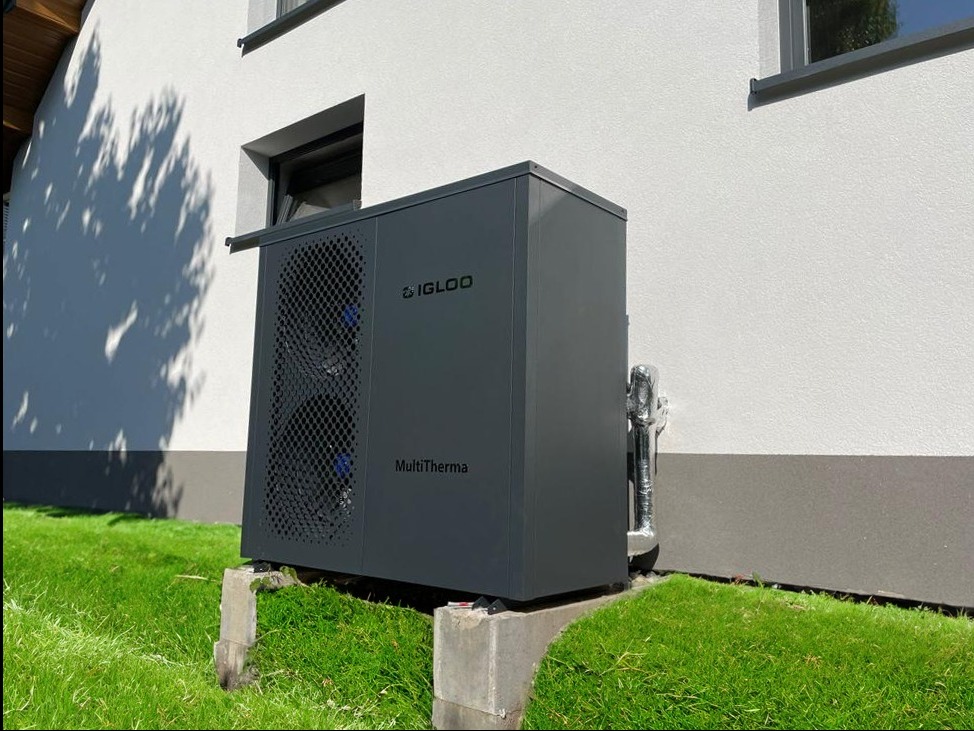 Szara pompa ciepła marki Igloo MultiTherma zainstalowana na betonowych podstawach przy ścianie budynku z białym tynkiem i szarym cokołem, otoczona zielonym trawnikiem.