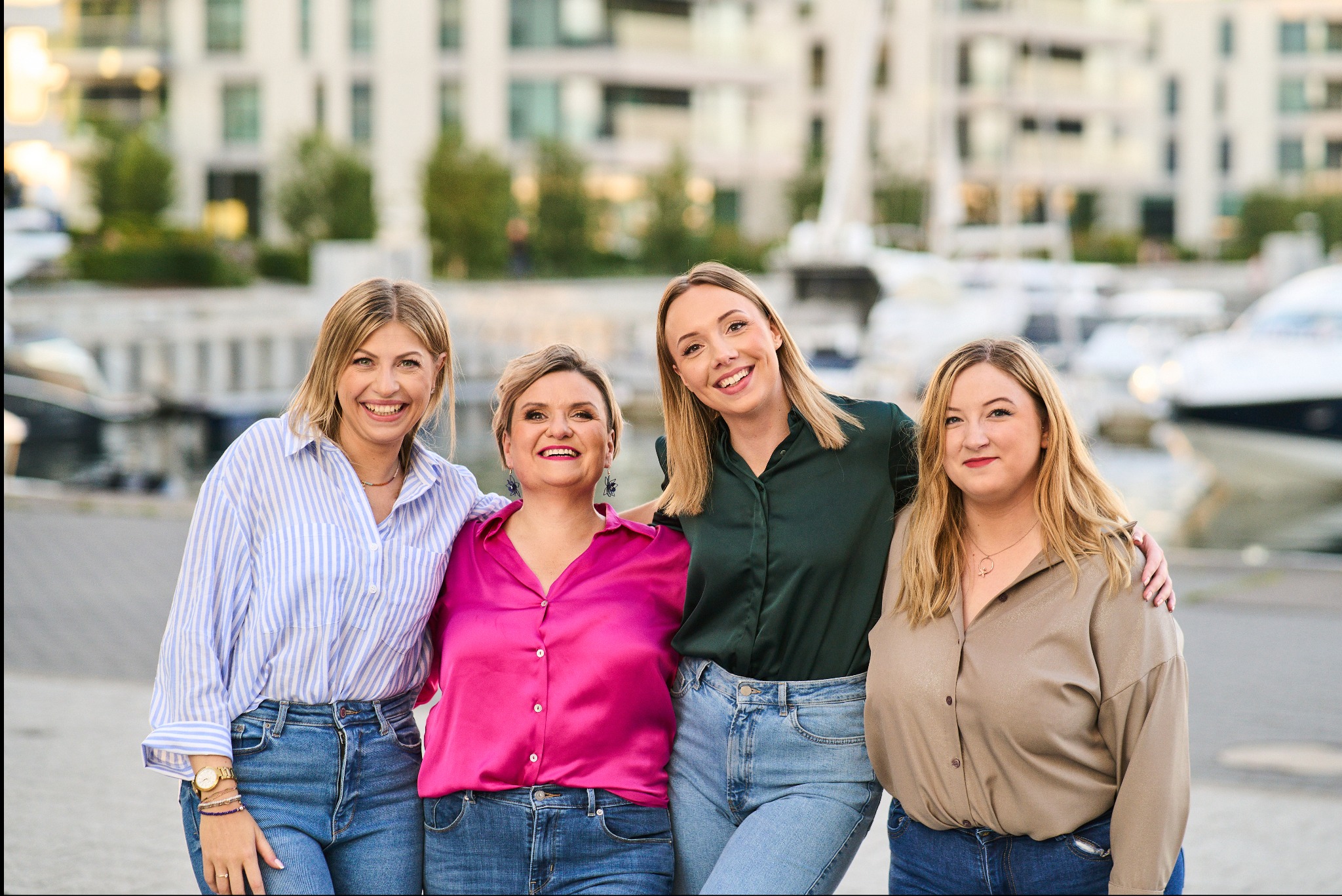 Cztery uśmiechnięte kobiety w różnym wieku, ubrane w bluzki i jeansy, stoją obok siebie na tle nowoczesnych budynków i zacumowanych łodzi w marinie.