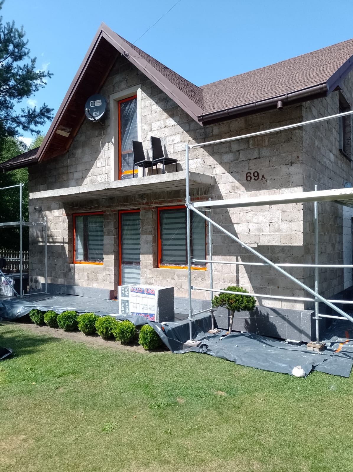 Dom jednorodzinny w trakcie prac, widoczne rusztowania, okna zabezpieczone folią, ocieplenie w blokach styropianowych, elewacja z szarego kamienia, numer domu 69A.