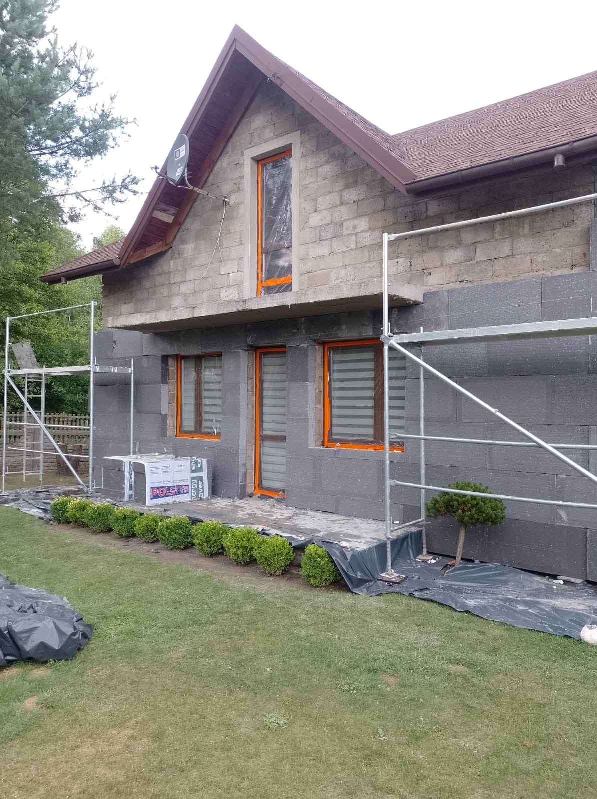 Dom w trakcie ocieplania styropianem grafitowym, widoczne rusztowania, okna zabezpieczone folią, paczki styropianu oraz małe krzewy przed budynkiem.