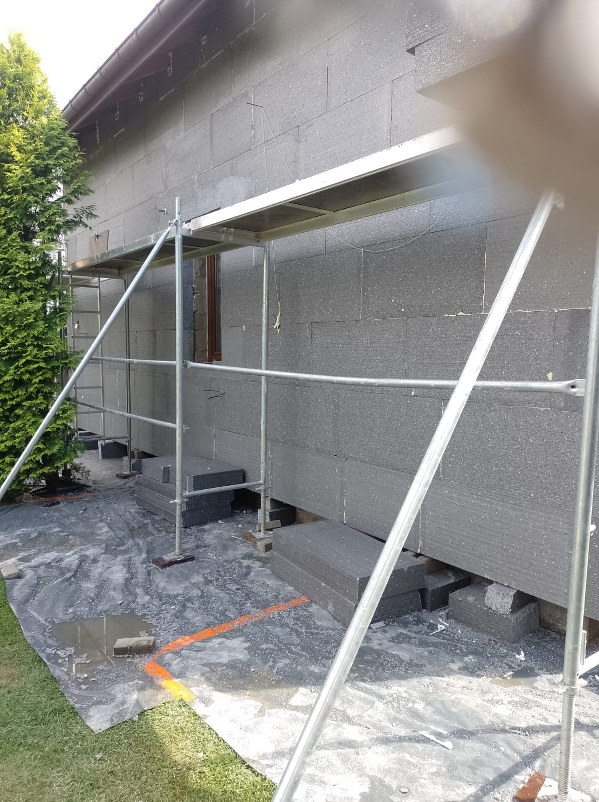 Ocieplanie elewacji budynku styropianem grafitowym, widoczne rusztowanie, zabezpieczona folią podstawa i fragment zielonego trawnika.
