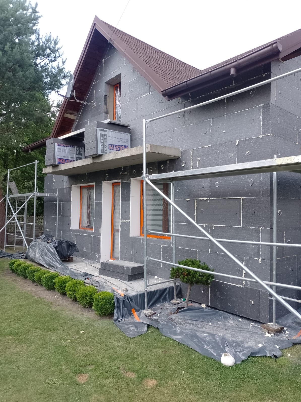 Dom jednorodzinny w trakcie ocieplania, widoczne szare płyty styropianowe na elewacji, rusztowanie, okna zabezpieczone folią, oraz ułożone kostki styropianowe i małe krzewy ozdobne wzdłuż ściany.