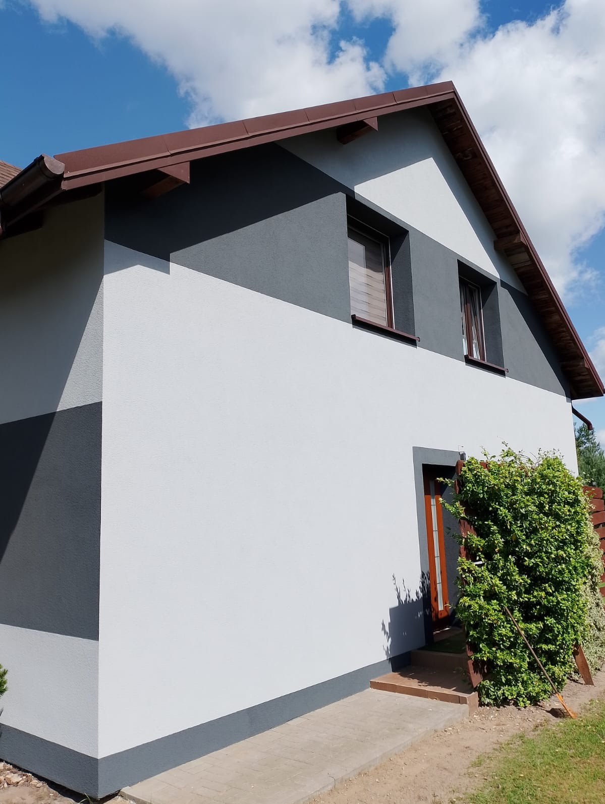 Dwukolorowa elewacja domu jednorodzinnego w odcieniach szarości i bieli, widok z boku, brązowy dach i stolarka okienna, bujna zieleń przy wejściu.