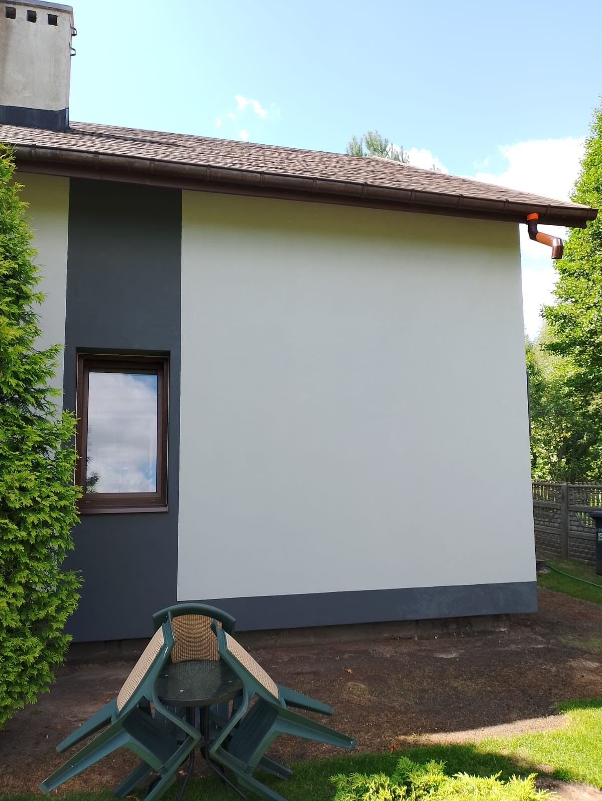 Świeżo pomalowana ściana budynku z dwukolorową elewacją, widoczne okno z brązową ramą, rynna i sterta plastikowych krzeseł ogrodowych