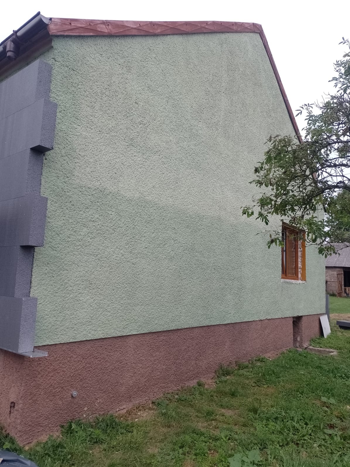 Fragment ściany domu z nałożonymi fragmentami szarego styropianu, widoczny zielony tynk strukturalny na pozostałej części elewacji, brązowa podmurówka i okno z drewnianą ramą.