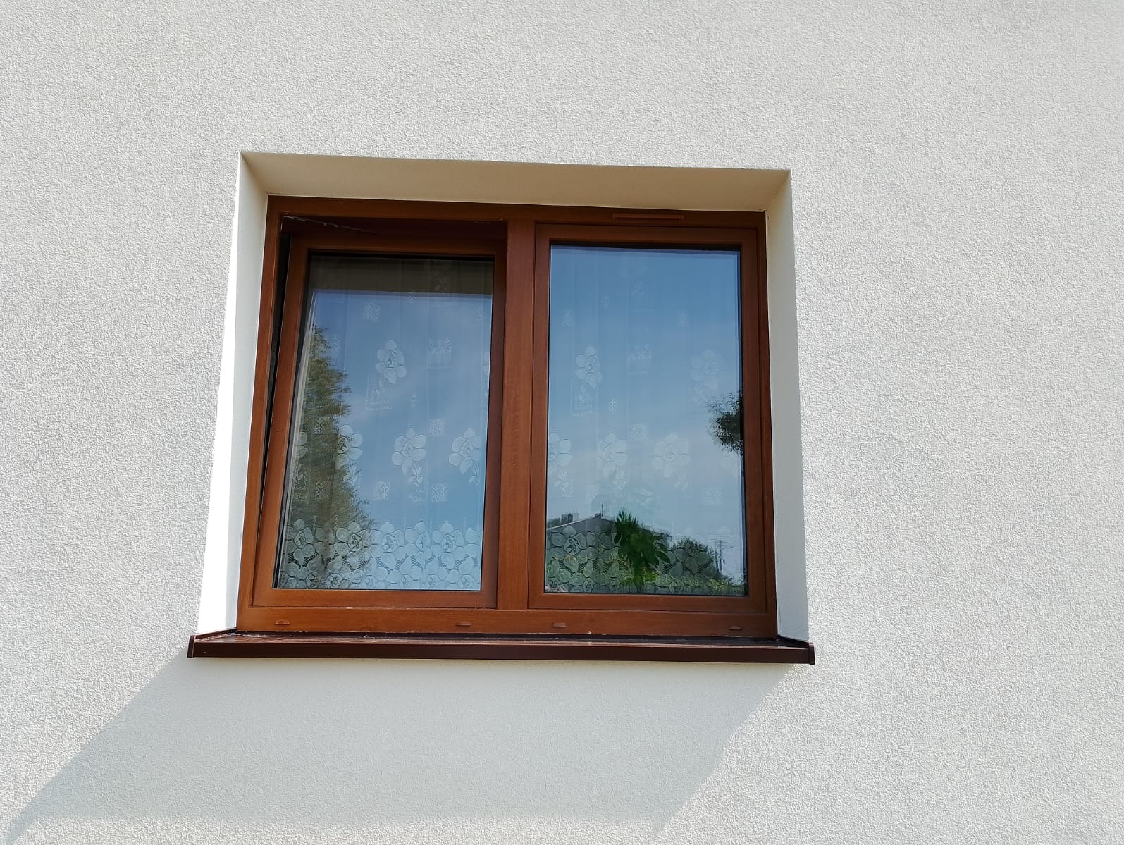 Dwukomorowe okno PCV w brązowej ramie, zamontowane w białej, teksturowanej elewacji budynku, z widocznym odbiciem nieba i drzew w szybach.