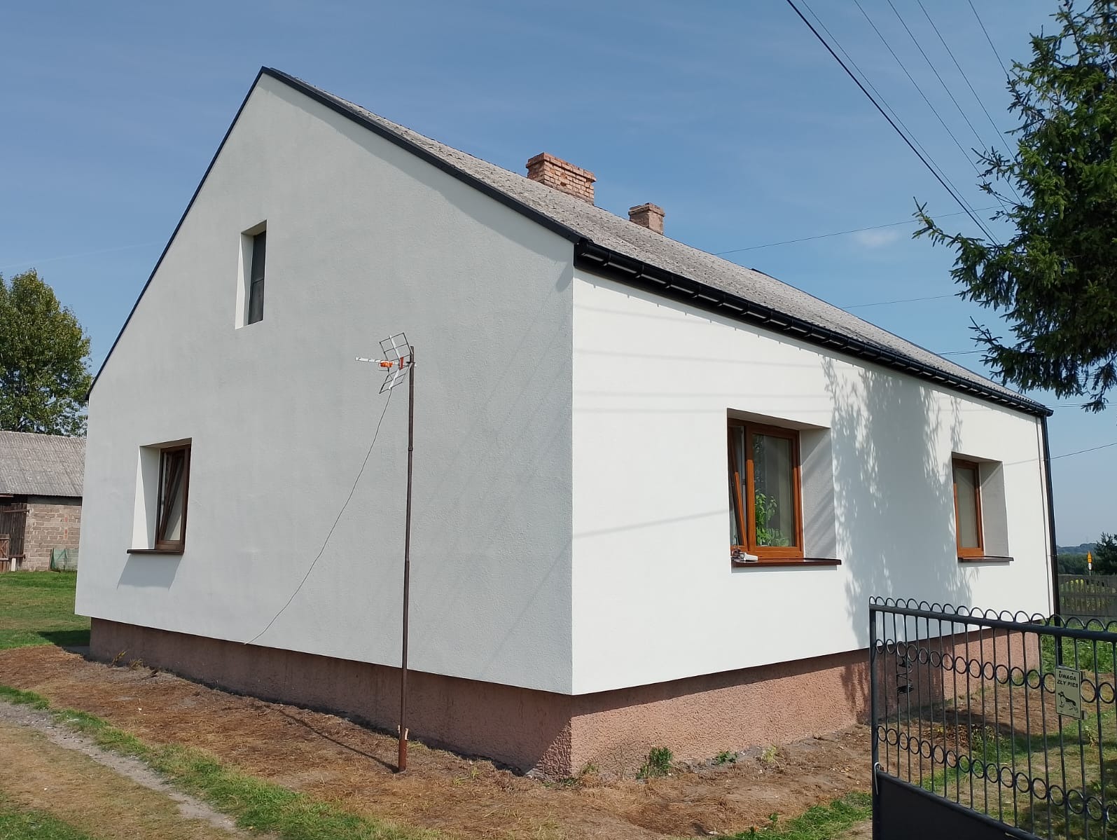 Boczny widok odnowionego domu jednorodzinnego z białą elewacją, brązowymi oknami i anteną telewizyjną na tle błękitnego nieba.