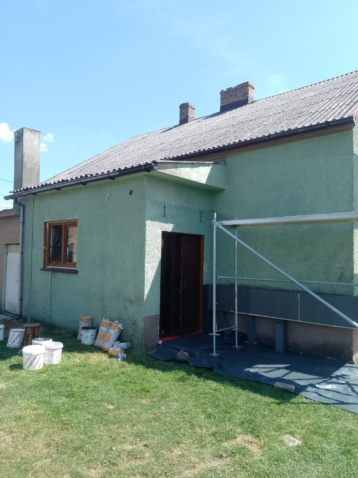 Dom jednorodzinny z ocieplonym fundamentem i elewacją w trakcie malowania, widoczne rusztowanie, wiaderka z farbą i worki z zaprawą na trawniku.