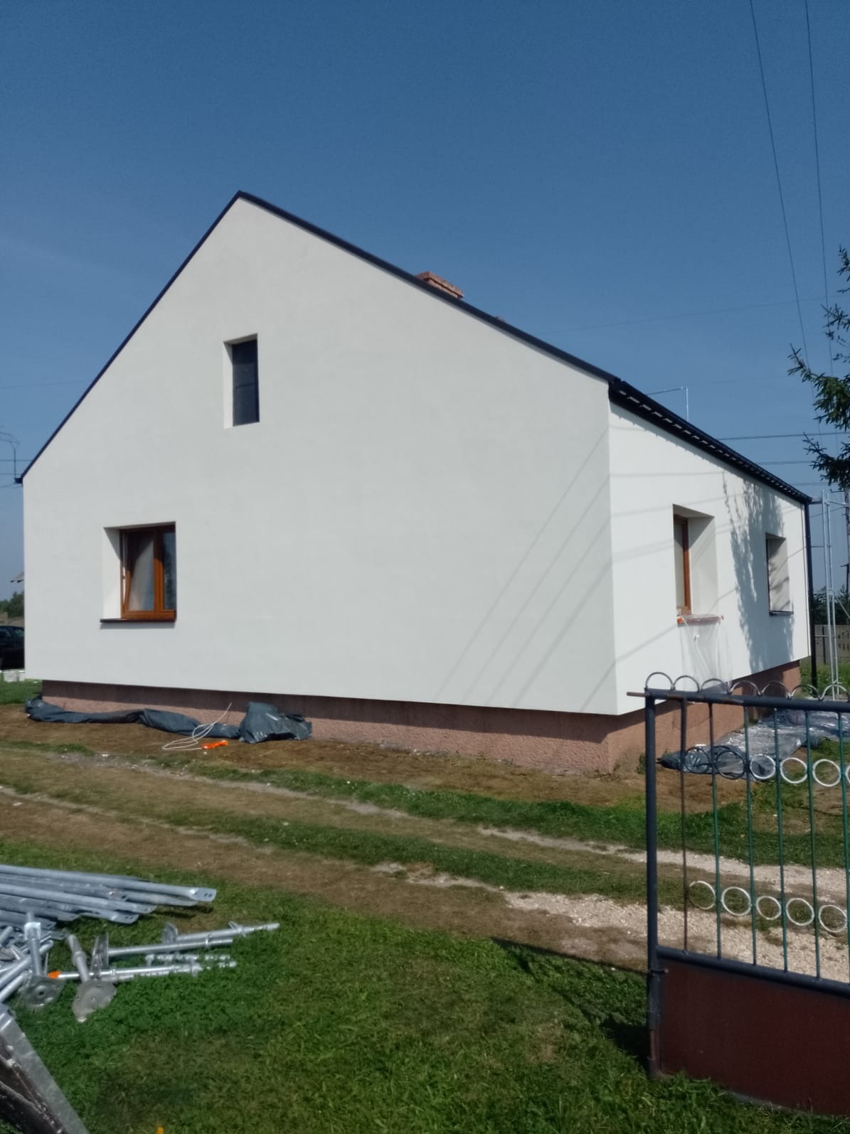 Świeżo otynkowany dom jednorodzinny z brązowymi oknami, widoczne rusztowania i folie zabezpieczające wokół budynku.