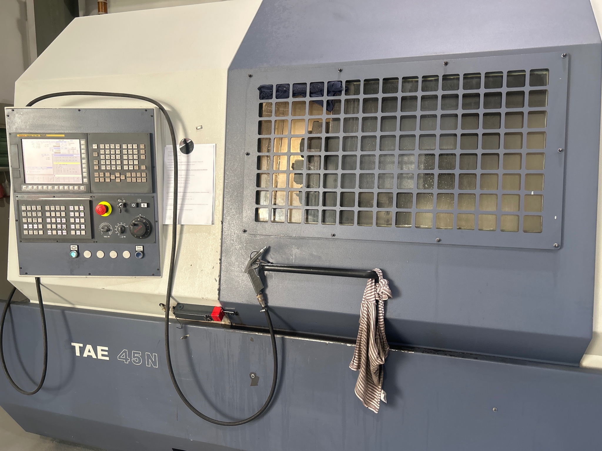 Szaro-biała maszyna CNC TAE 45N z panelem sterowania, osłoną z kratką i wiszącą szmatką w paski.