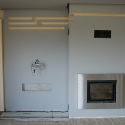 JARBUD - Ściana w trakcie wykańczania z wkładem kominkowym w metalowej obudowie, podświetlane wnęki i wystające przewody elektryczne.