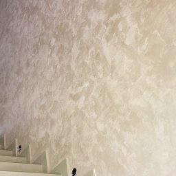 JARBUD - Ściana pokryta dekoracyjną gładzią w odcieniach beżu i bieli, widoczne schody z otworami na oświetlenie.