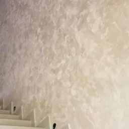 Ściana pokryta dekoracyjną gładzią w odcieniach beżu i bieli, widoczne schody z otworami na oświetlenie.