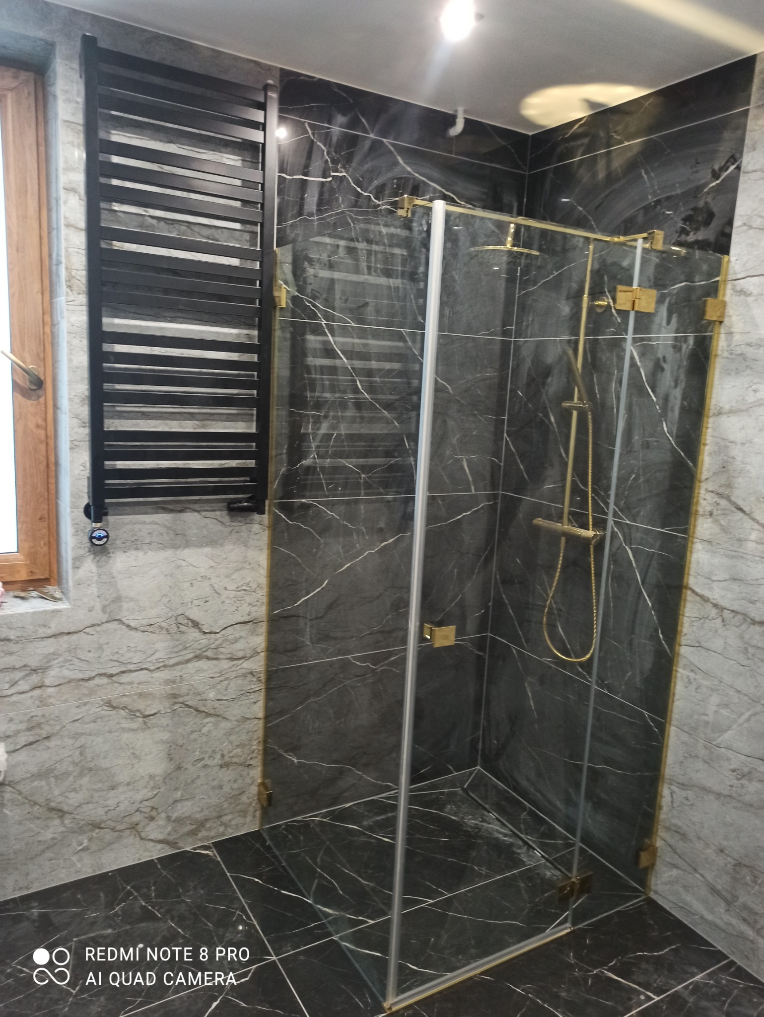 Nowoczesna kabina prysznicowa ze złotymi okuciami i czarną armaturą, wykończona marmurowymi płytkami w odcieniach szarości i czerni, z widocznym grzejnikiem drabinkowym.