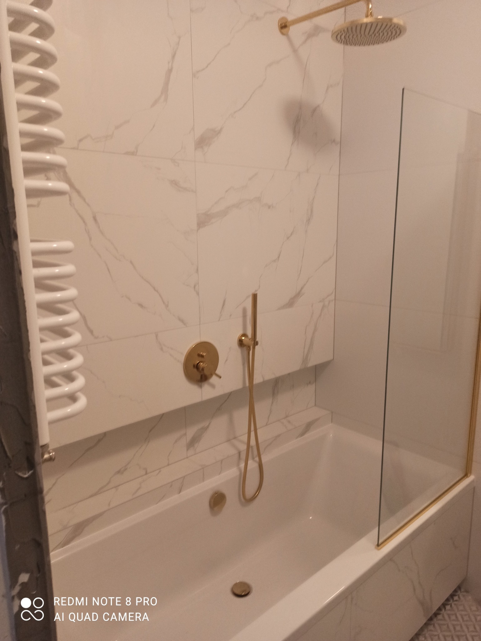 Nowoczesna łazienka z białą wanną obudowaną płytkami imitującymi marmur, złotą armaturą, szklanym parawanem nawannowym i białym grzejnikiem drabinkowym.