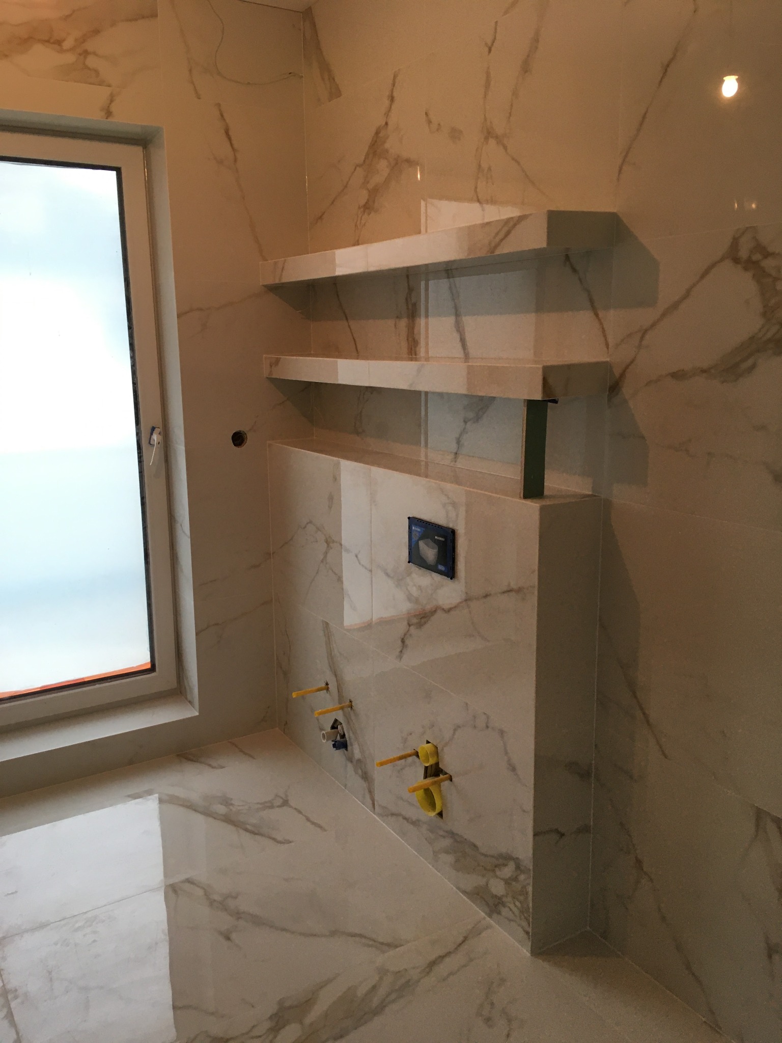 Wykończona łazienka z marmurowymi płytkami, podwieszana toaleta z przyciskiem spłukującym, dwie półki nad zabudową, widoczne żółte rurki instalacyjne, okno z widokiem na zewnątrz.