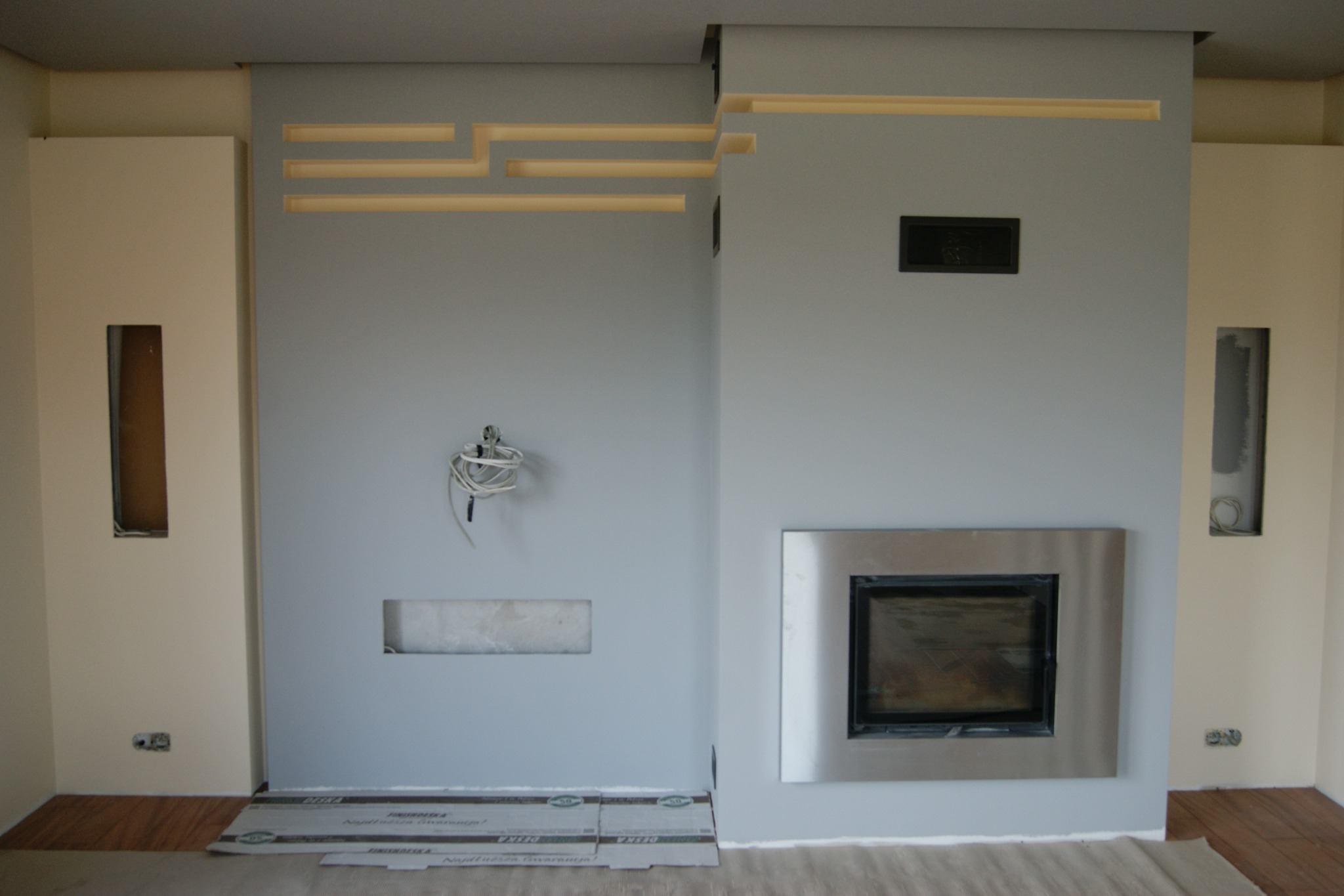 Ściana w trakcie wykańczania z wkładem kominkowym w metalowej obudowie, podświetlane wnęki i wystające przewody elektryczne.