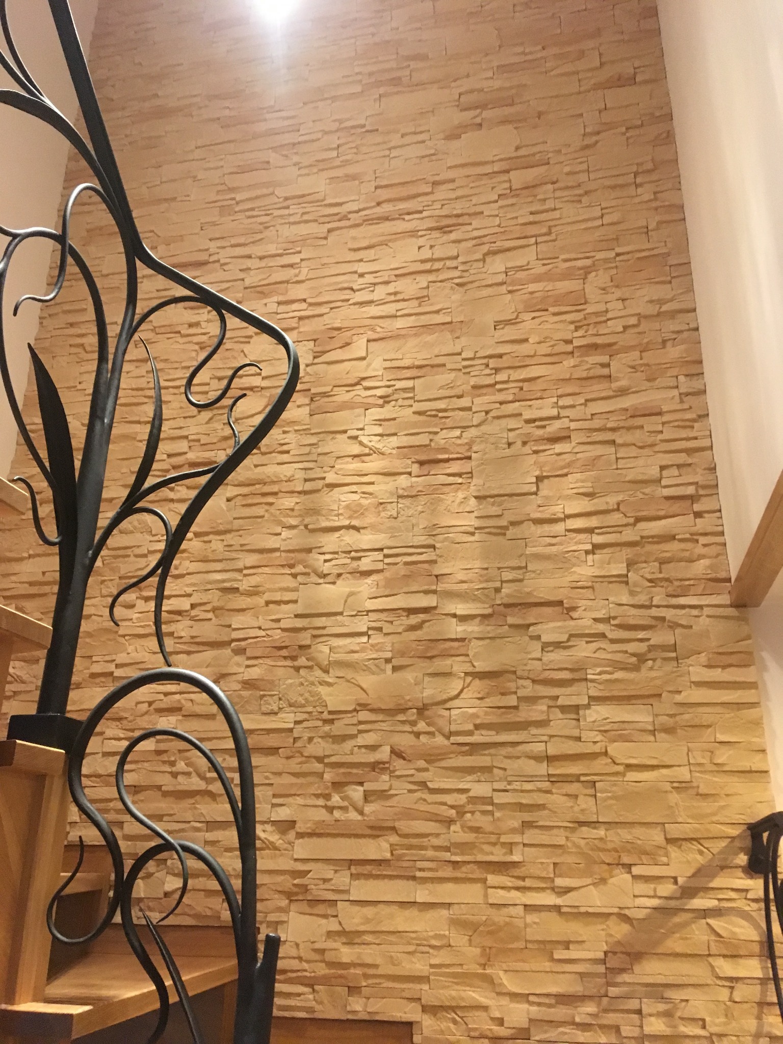 Ściana wykończona dekoracyjnym kamieniem w odcieniach beżu, widoczna z perspektywy schodów z ozdobną, kutą balustradą.
