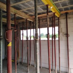 Daston - Wnętrze surowego domu w trakcie budowy, z widocznymi podporami stropowymi w kolorze czerwonym i żółtym, podtrzymującymi drewniane belki i betonowy strop.