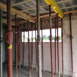 Wnętrze surowego domu w trakcie budowy, z widocznymi podporami stropowymi w kolorze czerwonym i żółtym, podtrzymującymi drewniane belki i betonowy strop.