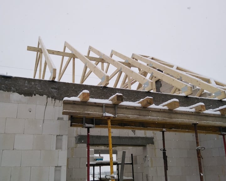 Wznoszenie drewnianej konstrukcji dachu na budynku z białych bloczków w zimowej scenerii, widoczne rusztowania i świeży śnieg.