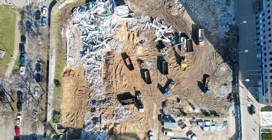 Widok z lotu ptaka na plac budowy z gruzem, wykopami, ciężarówkami i koparką, obok parkingu i budynku.