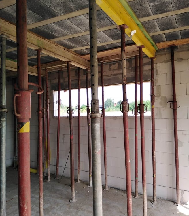 Wnętrze surowego domu w trakcie budowy, z widocznymi podporami stropowymi w kolorze czerwonym i żółtym, podtrzymującymi drewniane belki i betonowy strop.