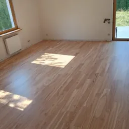 Jasne panele podłogowe imitujące drewno w pustym pokoju z oknem i drzwiami balkonowymi, widoczne ślady po listwach przypodłogowych.