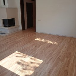 Mariusz Wiórek - Jasne, drewniane panele podłogowe ułożone w pustym pokoju z kominkiem i otwartymi drzwiami, widoczne ślady po listwach przypodłogowych.