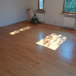 Pokój z wycyklinowaną, błyszczącą podłogą drewnianą, widoczna maszyna do cyklinowania Lägler i wąż od odkurzacza, ślady po listwach przypodłogowych na ścianach, wpadające promienie słoneczne.