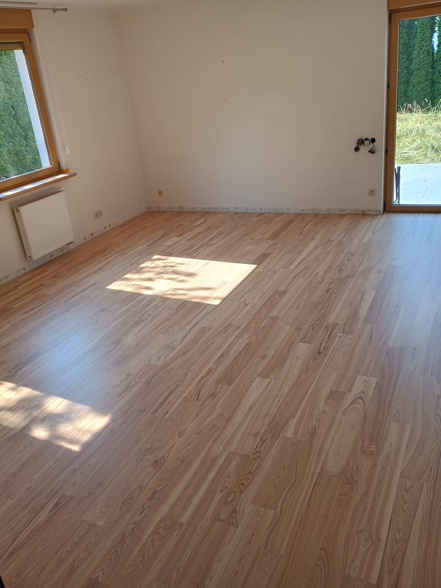 Jasne panele podłogowe imitujące drewno w pustym pokoju z oknem i drzwiami balkonowymi, widoczne ślady po listwach przypodłogowych.