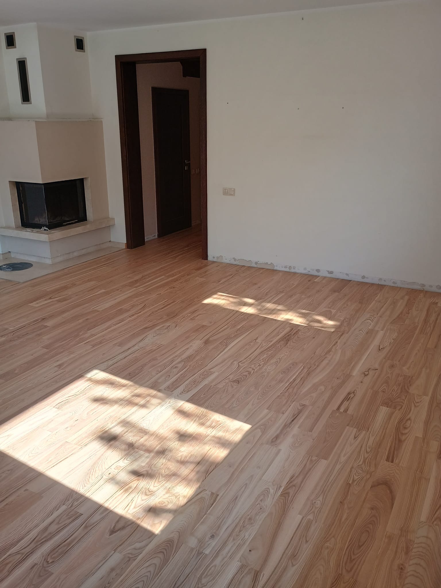 Jasne, drewniane panele podłogowe ułożone w pustym pokoju z kominkiem i otwartymi drzwiami, widoczne ślady po listwach przypodłogowych.