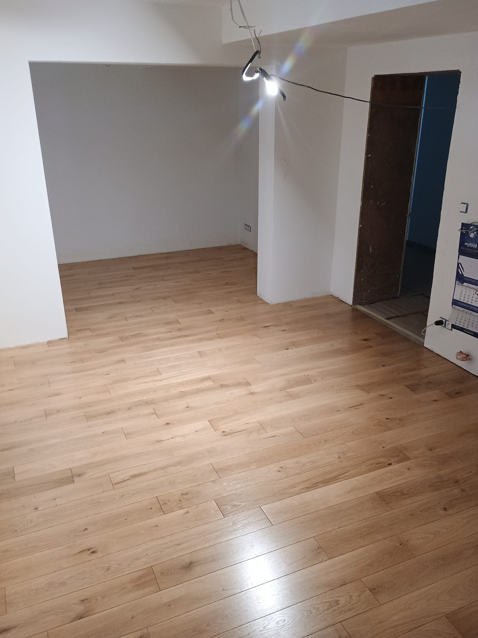 Jasne panele podłogowe ułożone w nowym pomieszczeniu z surowymi ścianami i wiszącą lampą budowlaną, widoczne niedokończone futryny drzwi.