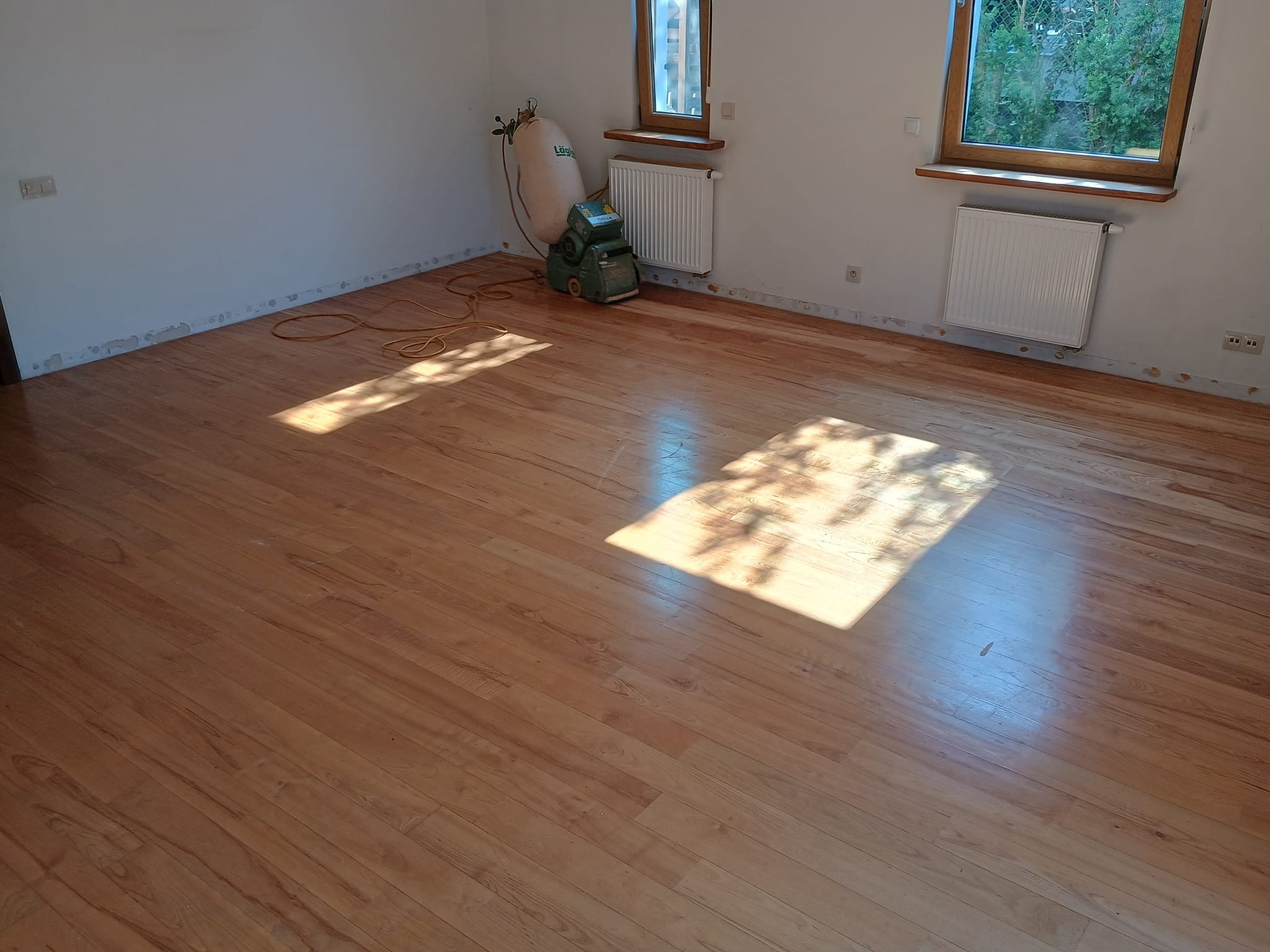 Pokój z wycyklinowaną, błyszczącą podłogą drewnianą, widoczna maszyna do cyklinowania Lägler i wąż od odkurzacza, ślady po listwach przypodłogowych na ścianach, wpadające promienie słoneczne.