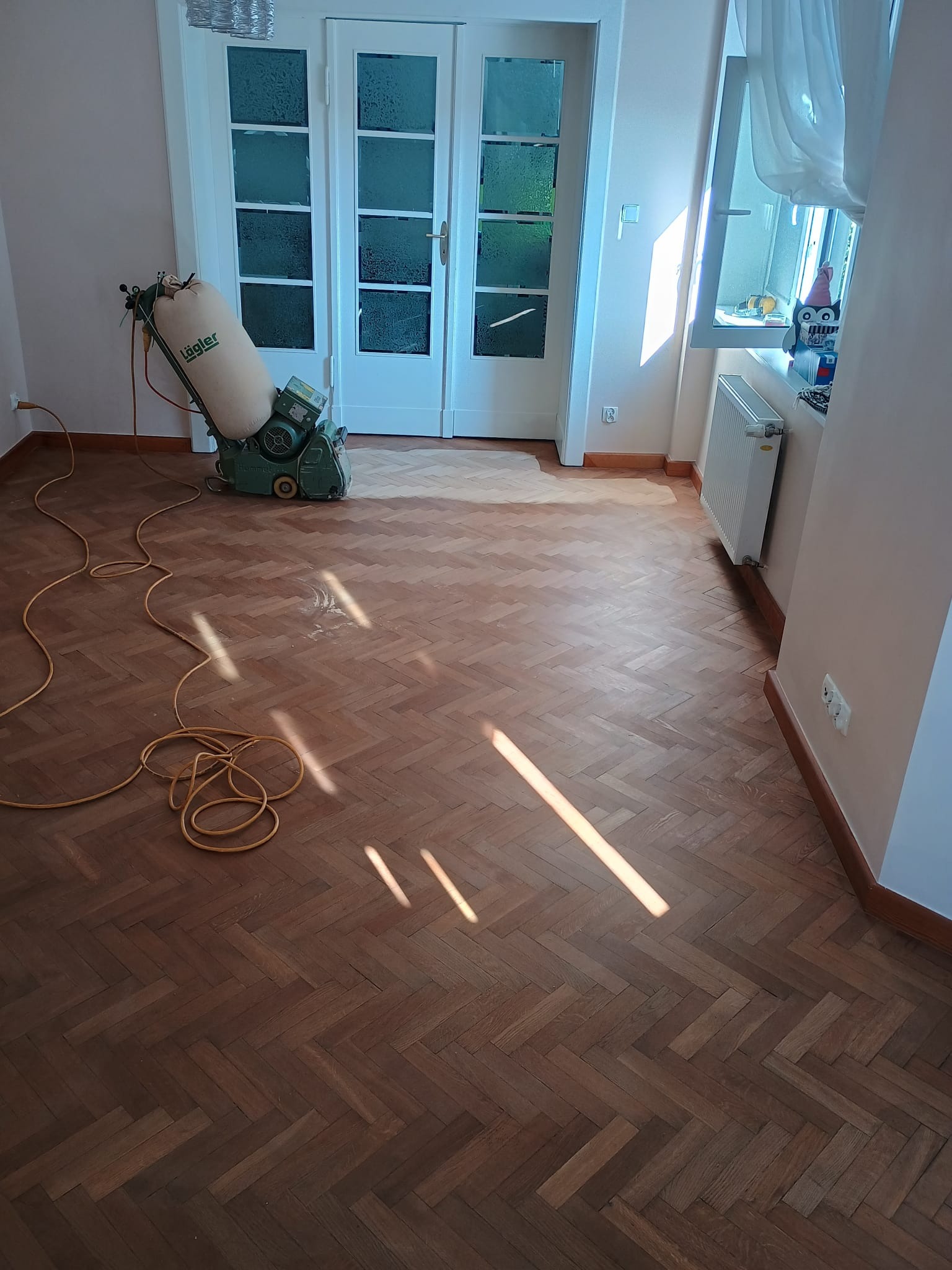 Parkiet ułożony w jodełkę, częściowo wycyklinowany, z widoczną maszyną Lägler Hummel i kablem zasilającym na podłodze, w tle białe dwuskrzydłowe drzwi i okno z grzejnikiem.