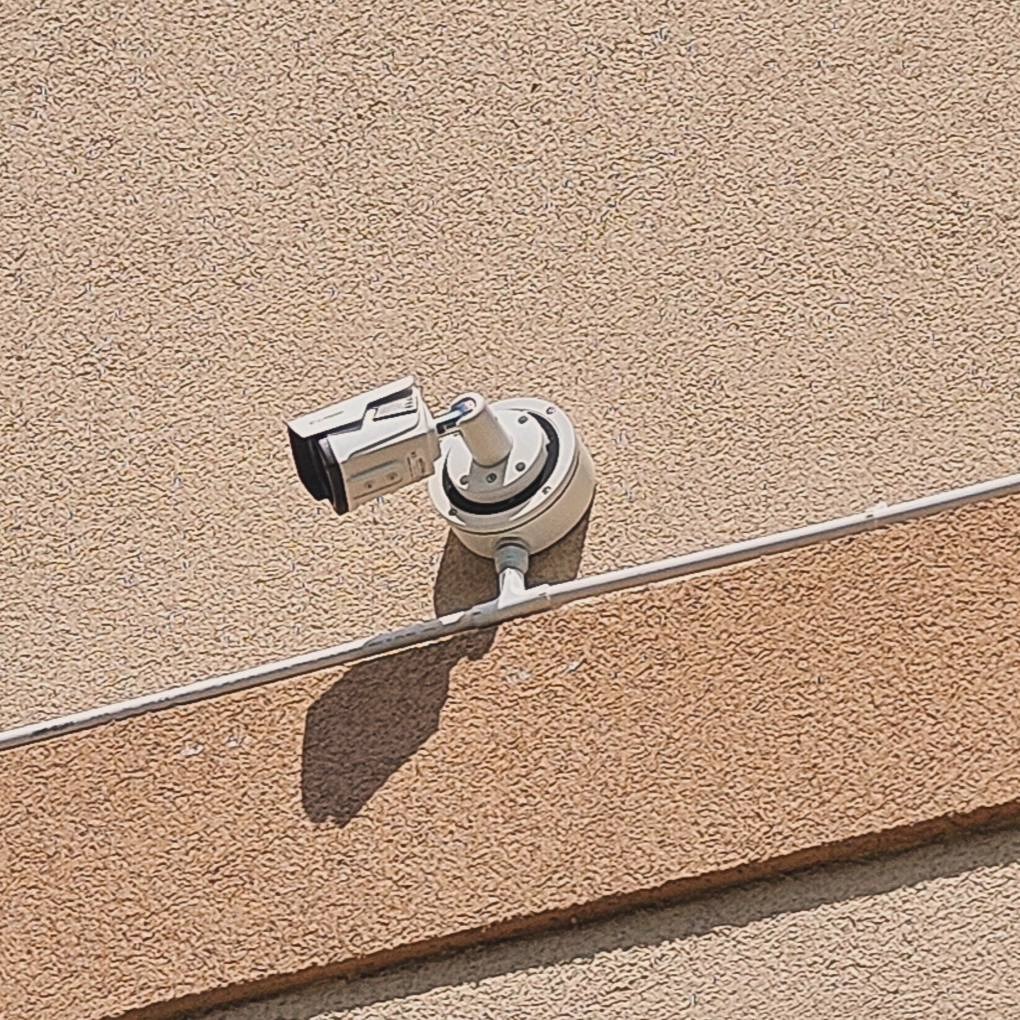 Zewnętrzna kamera monitoringu CCTV zamontowana na teksturowanej ścianie budynku, widoczne okablowanie i cień kamery w słoneczny dzień.