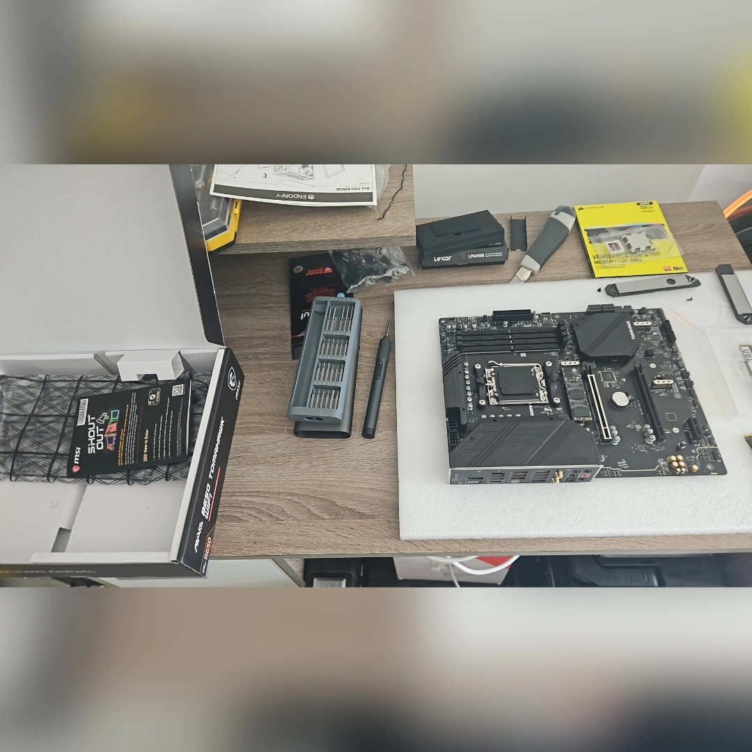 Płyta główna MSI z serii MAG B650 Tomahawk WiFi, wyjęta z pudełka, obok zestawu precyzyjnych wkrętaków, pamięci RAM Lexar i Corsair oraz instrukcji obsługi, na stole o drewnianej fakturze.