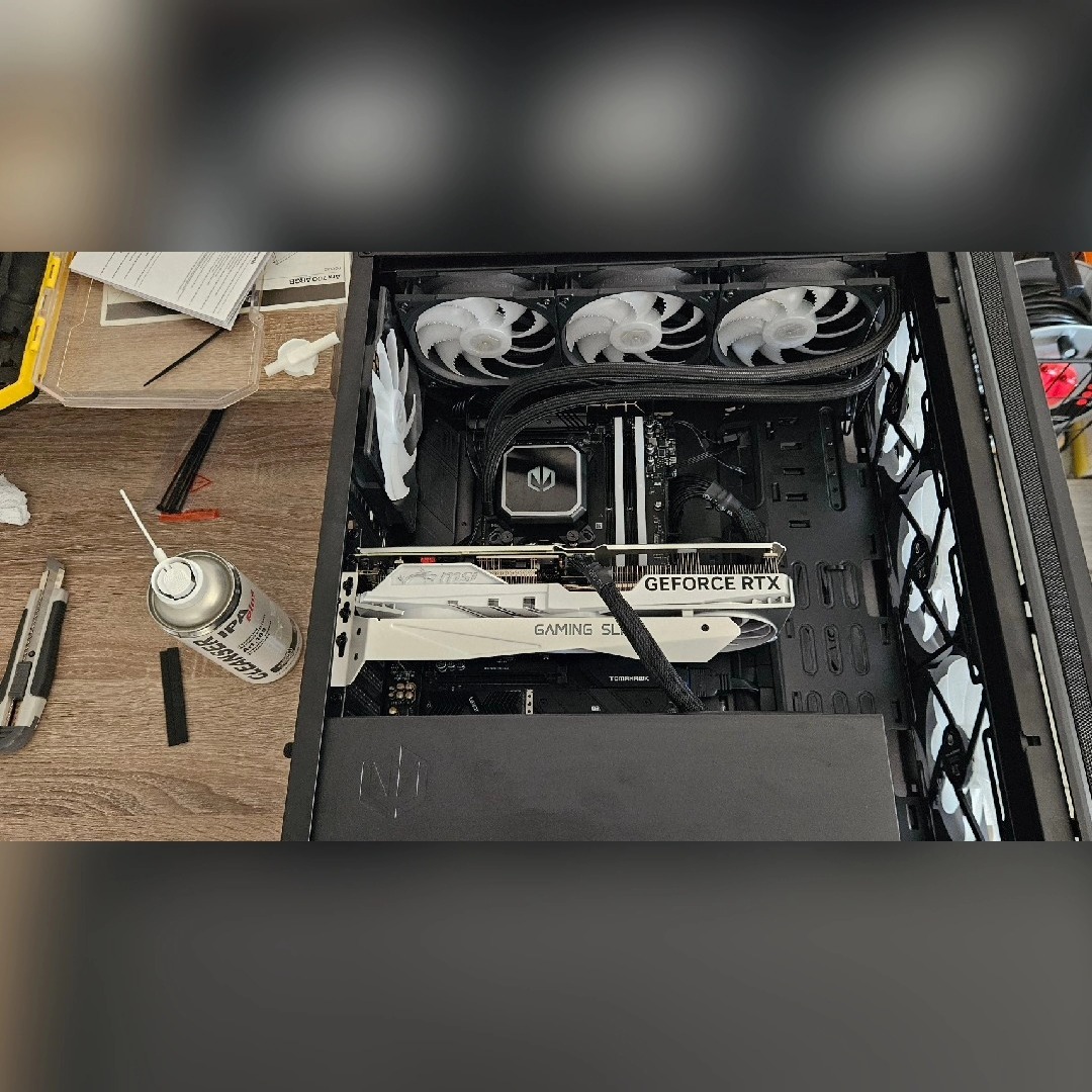 Wnętrze komputera stacjonarnego z białą kartą graficzną GeForce RTX, chłodzeniem wodnym i trzema wentylatorami, obok leży puszka sprężonego powietrza i nożyk introligatorski.