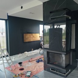 GLAZ TECH SZYSZKA RYSZARD - Wnętrze w trakcie remontu z czarnymi ścianami, marmurowym kominkiem i rozłożonymi foliami ochronnymi na podłodze. Malarz w tle maluje ścianę na złoty kolor.