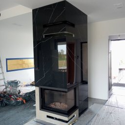 GLAZ TECH SZYSZKA RYSZARD - Wnętrze w trakcie prac wykończeniowych, widoczny nowoczesny kominek obłożony czarnym marmurem z białymi żyłami, zabezpieczona folią podłoga, drabina i odkurzacz przemysłowy w tle.