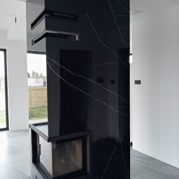 GLAZ TECH SZYSZKA RYSZARD - Nowoczesny kominek obłożony czarnym marmurem z białymi żyłami, widoczny w surowym wnętrzu z szarą podłogą i białymi ścianami, z widokiem na budowę za oknem.