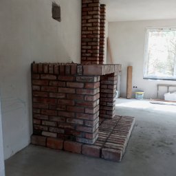 GLAZ TECH SZYSZKA RYSZARD - Murowany kominek z cegły w surowym stanie, widoczne niedoskonałości spoin, ściany w trakcie wykończenia, otwór w ścianie, okno z widokiem na drzewa w tle.