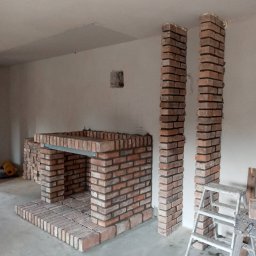GLAZ TECH SZYSZKA RYSZARD - Wnętrze w trakcie budowy: murowany kominek z czerwonej cegły, wsparty na metalowej belce, z dwoma kolumnami z cegły po bokach, drabina i narzędzia w tle, surowy stan wykończenia.