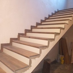 GLAZ TECH SZYSZKA RYSZARD - Drewniane schody z białymi podstopnicami w trakcie wykończenia, widoczne ślady zaprawy i niedomalowane fragmenty, w tle worki i deski.