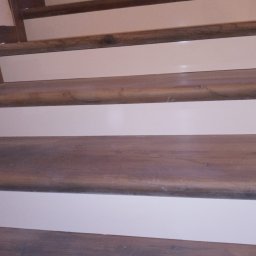 GLAZ TECH SZYSZKA RYSZARD - Bieg schodów z drewnianymi stopnicami o zaokrąglonych krawędziach i białymi podstopnicami, widok z dołu.