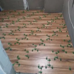 Proces układania podłogi z płytek imitujących drewno, z użyciem zielonych krzyżyków dystansowych, na nierównej powierzchni w trakcie remontu.