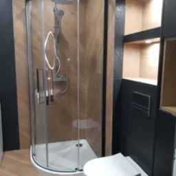 Nowoczesna łazienka z kabiną prysznicową narożną z czarną ramą, wiszącą toaletą, drewnopodobnymi płytkami ułożonymi w jodełkę oraz czarnymi płytkami na ścianie z wnękami oświetlonymi od góry.