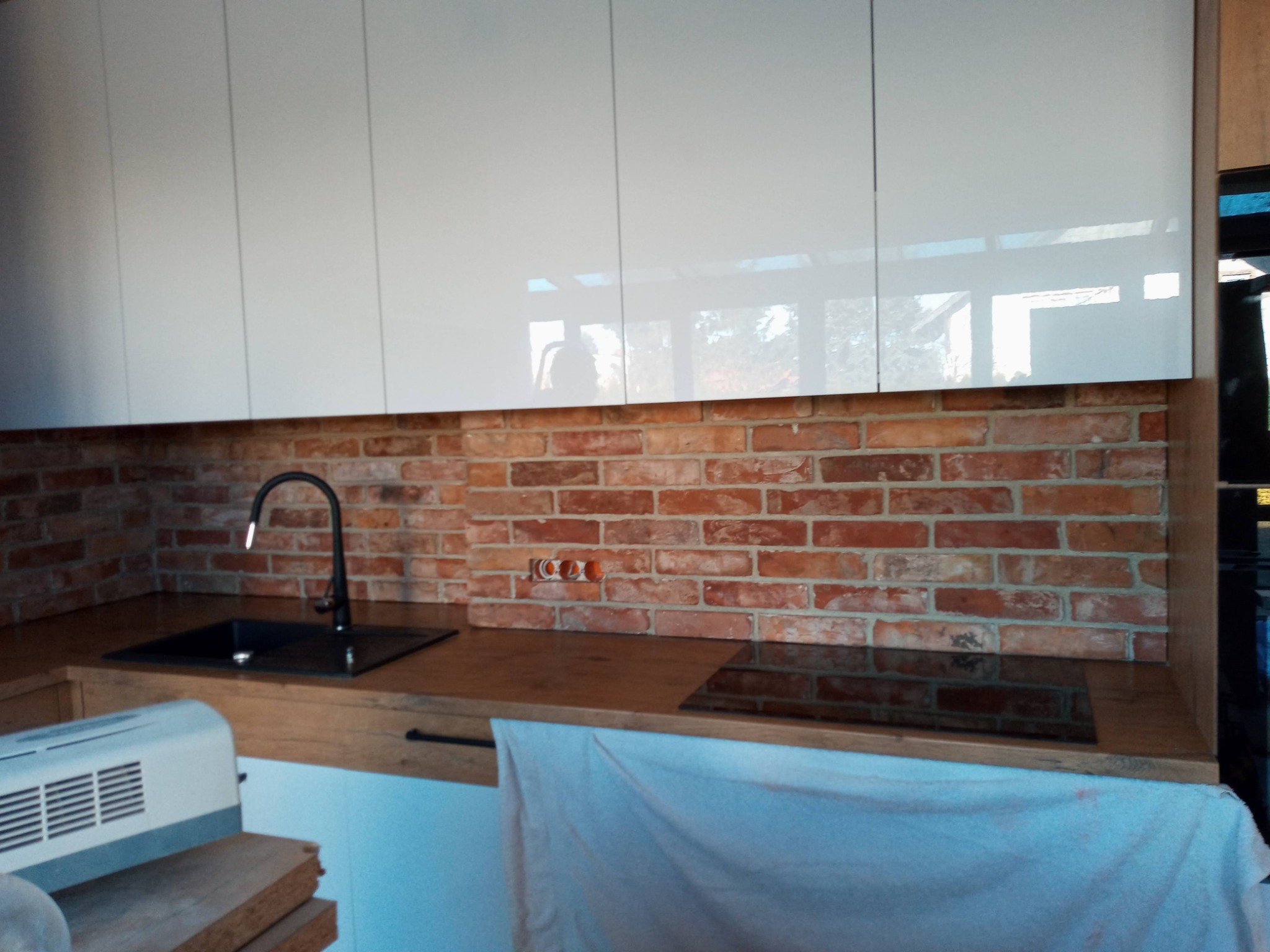 Częściowy widok kuchni w trakcie prac wykończeniowych: białe, błyszczące szafki wiszące nad blatem z ciemnego drewna i ceglaną ścianą, czarny zlew i płyta indukcyjna, widoczne gniazdka elektryczne,...