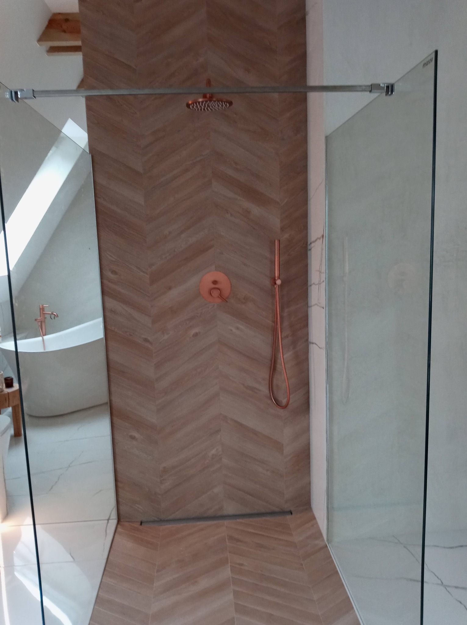 Nowoczesna łazienka z kabiną prysznicową typu walk-in, wykończona płytkami w jodełkę w odcieniach brązu, z armaturą w kolorze miedzi oraz widoczną w lustrze wolnostojącą wanną.